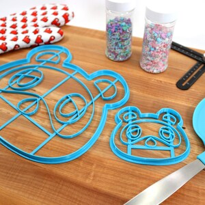 May include: Two blue plastic cookie cutters shaped like a cartoon animal face, one larger than the other, on a wooden surface. There are two jars of sprinkles, a rolling pin, a spatula, and a ruler in the background.