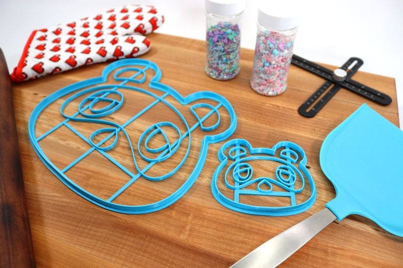 May include: Two blue plastic cookie cutters shaped like a cartoon animal face, one larger than the other, on a wooden surface. There are two jars of sprinkles, a rolling pin, a spatula, and a ruler in the background.