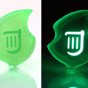 May include: A green, shield-shaped emblem with a white, stylized symbol. The object glows in the dark, with the symbol illuminated. The design is reminiscent of a video game emblem.