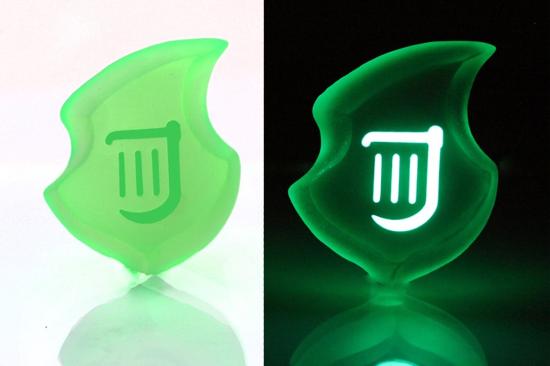 May include: A green, shield-shaped emblem with a white, stylized symbol. The object glows in the dark, with the symbol illuminated. The design is reminiscent of a video game emblem.