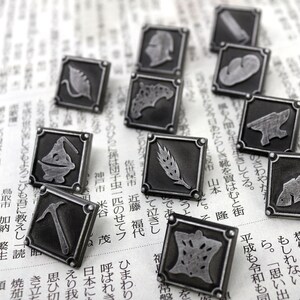May include: A collection of 11 silver-toned metal pins with various designs, including a fish, a wheat stalk, a pickaxe, a cauldron, a heart, a fish, a mushroom, a leaf, a helmet, a horseshoe, and a bird. The pins are arranged on a newspaper background.
