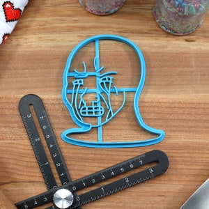 Op de afbeelding: Een blauwe plastic koekjesvormer in de vorm van een menselijk gezicht met een rasterpatroon op het gezicht.