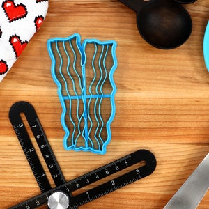 May include: A blue plastic bacon-shaped cookie cutter sits on a wooden surface, next to a heart-patterned oven mitt, black measuring spoons, a blue spatula, and a black angle finder tool. The cutter has wavy lines to mimic bacon strips.