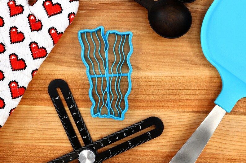 May include: A blue plastic bacon-shaped cookie cutter sits on a wooden surface, next to a heart-patterned oven mitt, black measuring spoons, a blue spatula, and a black angle finder tool. The cutter has wavy lines to mimic bacon strips.