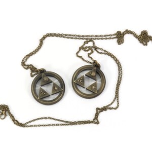 Puede incluir: Dos collares de color dorado con un colgante de Trifuerza. Cada colgante presenta un dise&ntilde;o triangular con tres tri&aacute;ngulos m&aacute;s peque&ntilde;os en el interior, que representan las tres diosas del poder, la sabidur&iacute;a y el valor.