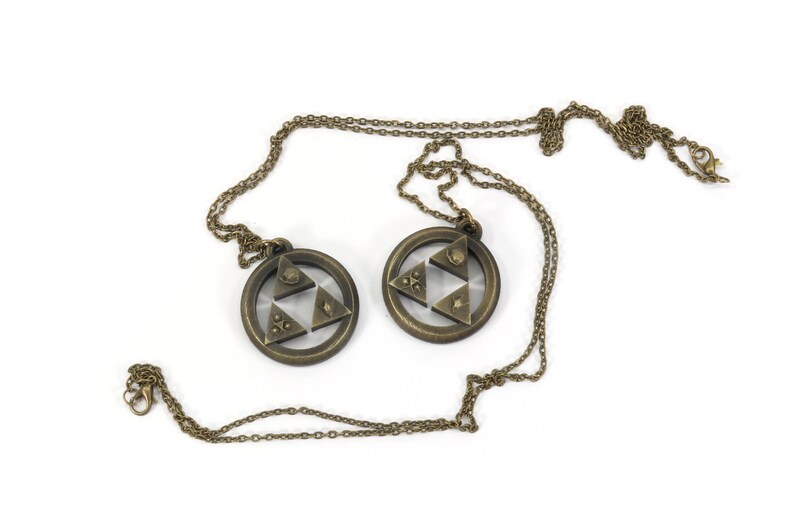 Puede incluir: Dos collares de color dorado con un colgante de Trifuerza. Cada colgante presenta un dise&ntilde;o triangular con tres tri&aacute;ngulos m&aacute;s peque&ntilde;os en el interior, que representan las tres diosas del poder, la sabidur&iacute;a y el valor.