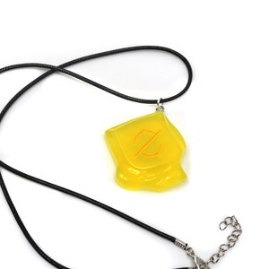 May include: A yellow pendant necklace with a black cord. The pendant is a translucent, irregularly shaped square with an orange symbol. The necklace has a silver-colored clasp.