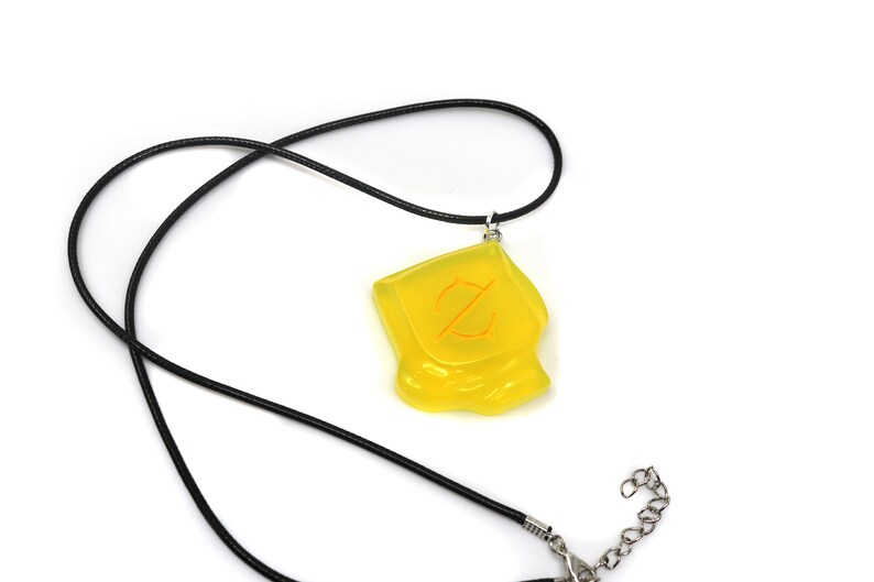May include: A yellow pendant necklace with a black cord. The pendant is a translucent, irregularly shaped square with an orange symbol. The necklace has a silver-colored clasp.