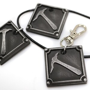 May include: Three square pendants with a pickaxe design, connected by a black cord and a silver clasp. The pendants are dark gray with a metallic pickaxe in the center. The design is a close-up shot on a white background.