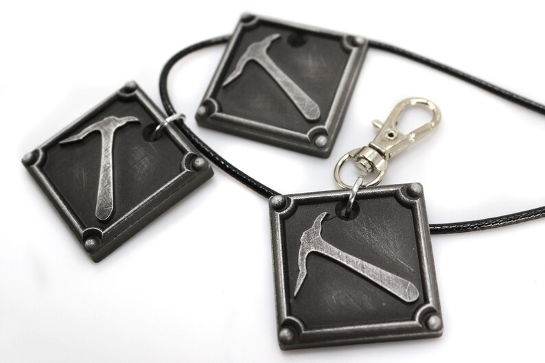 May include: Three square pendants with a pickaxe design, connected by a black cord and a silver clasp. The pendants are dark gray with a metallic pickaxe in the center. The design is a close-up shot on a white background.
