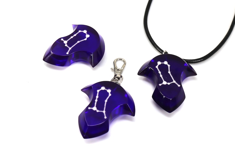 May include: Three blue glass pendants with a white Gemini constellation design. One pendant is on a black cord necklace, one is on a silver key chain, and one is laying on a white surface.