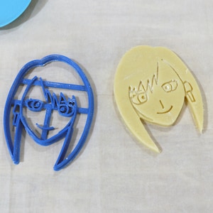 May include: Two cookie cutters, one blue and one yellow, shaped like a person's face with hair. The blue cutter has a more detailed design with eyes, a nose, and a mouth.