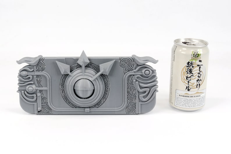 May include: A gray 3D printed case for a Nintendo Switch console. The case features an intricate design with a circular element in the center and a raised, textured pattern. The case is about the size of a standard can of soda.