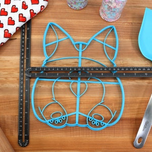May include: A blue plastic cookie cutter shaped like a dog, with a heart-shaped nose and paw prints on the bottom. The cutter is on a wooden surface with a ruler and a spatula.