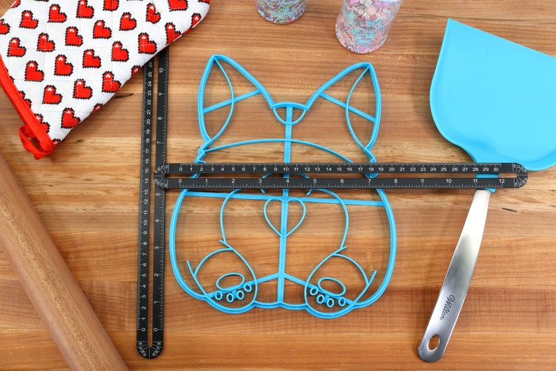 May include: A blue plastic cookie cutter shaped like a dog, with a heart-shaped nose and paw prints on the bottom. The cutter is on a wooden surface with a ruler and a spatula.