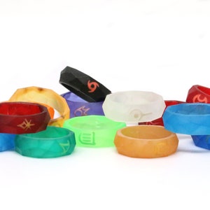 Pu&ograve; includere: Una collezione di 12 anelli in silicone colorati con diversi simboli e disegni. Gli anelli sono impilati e disposti a semicerchio su una superficie bianca.
