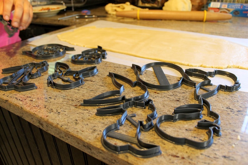 May include: A set of eight grey plastic cookie cutters in various shapes, including a circle, a star, a flower, and a leaf. The cutters are arranged on a brown work surface.