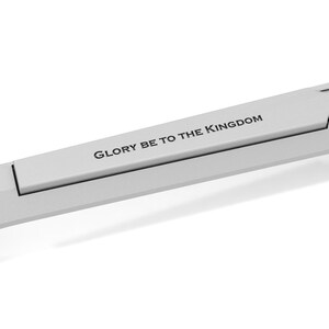 May include: A light gray, metallic object with the text "GLORY BE TO THE KINGDOM" printed on it. The object has a unique shape with a rectangular section and curved edges, set against a white background.