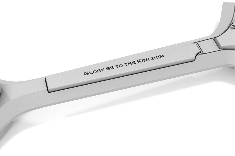 May include: A light gray, metallic object with the text "GLORY BE TO THE KINGDOM" printed on it. The object has a unique shape with a rectangular section and curved edges, set against a white background.