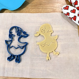 May include: A blue plastic cookie cutter and a yellow cookie in the shape of a chick on a white parchment paper surface.