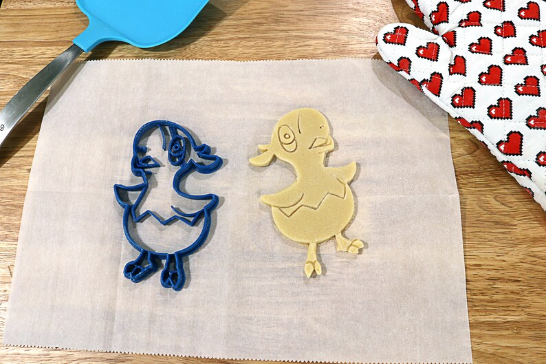 May include: A blue plastic cookie cutter and a yellow cookie in the shape of a chick on a white parchment paper surface.