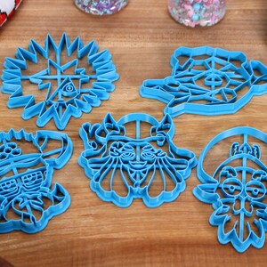 May include: Set of six blue plastic cookie cutters featuring cartoon characters with various facial expressions. The cutters are on a wooden surface.