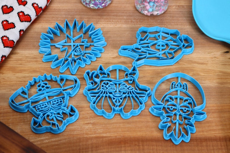 May include: Set of six blue plastic cookie cutters featuring cartoon characters with various facial expressions. The cutters are on a wooden surface.