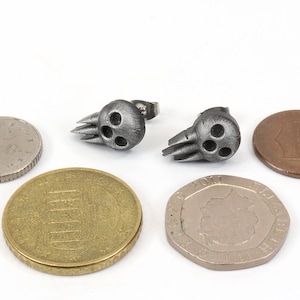 Peut inclure: Une paire de boucles d'oreilles en forme de cr&acirc;ne argent&eacute;es avec une finition textur&eacute;e. Les boucles d'oreilles sont pr&eacute;sent&eacute;es &agrave; c&ocirc;t&eacute; de plusieurs pi&egrave;ces de monnaie pour comparaison de taille.