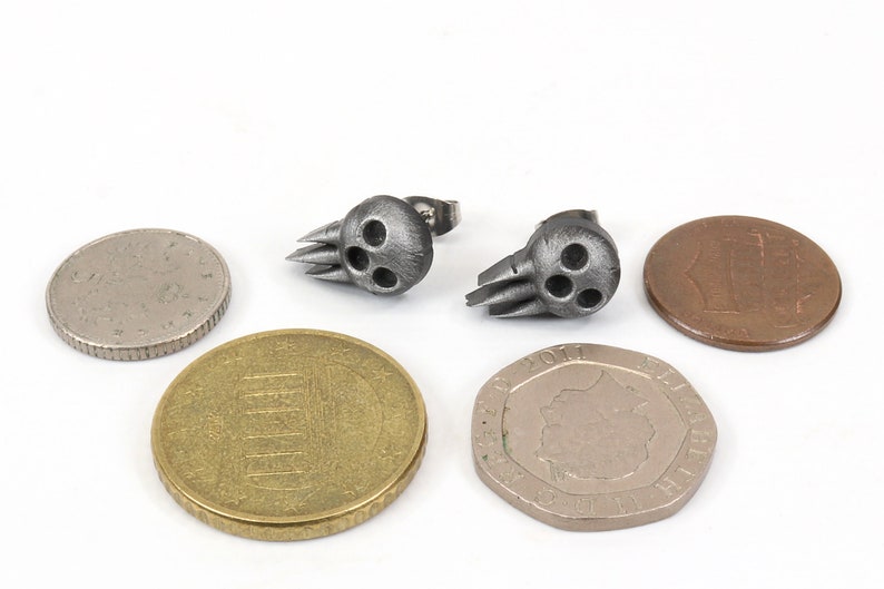 Peut inclure: Une paire de boucles d'oreilles en forme de cr&acirc;ne argent&eacute;es avec une finition textur&eacute;e. Les boucles d'oreilles sont pr&eacute;sent&eacute;es &agrave; c&ocirc;t&eacute; de plusieurs pi&egrave;ces de monnaie pour comparaison de taille.