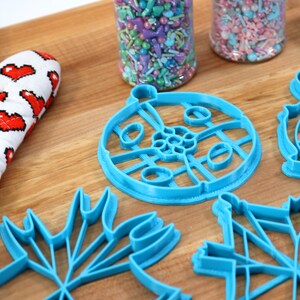 May include: A set of five blue plastic cookie cutters shaped like a snowflake, a Christmas tree, a star, a circle with a design, and a globe. The cutters are on a wooden surface.
