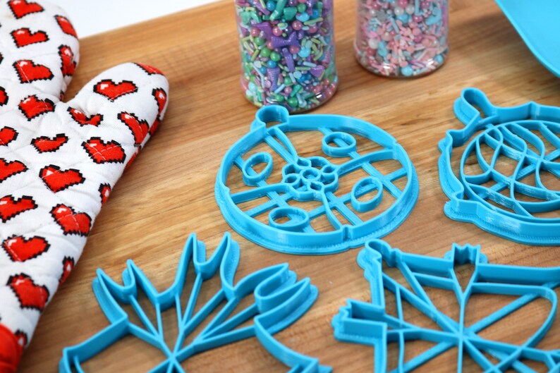 May include: A set of five blue plastic cookie cutters shaped like a snowflake, a Christmas tree, a star, a circle with a design, and a globe. The cutters are on a wooden surface.