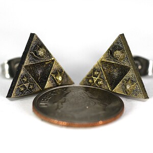 May include: A pair of gold-toned triangle earrings with a black and gold design. The earrings are about the size of a US penny.