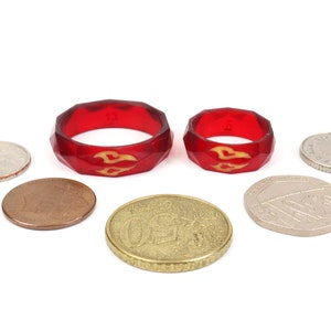 May include: Two red silicone rings with a gold flame design. The rings are different sizes and are sitting on a white surface with several coins.