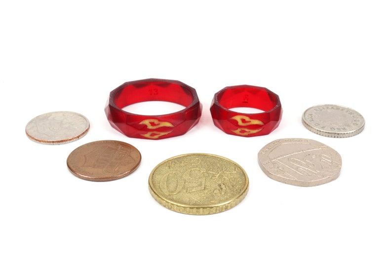 May include: Two red silicone rings with a gold flame design. The rings are different sizes and are sitting on a white surface with several coins.