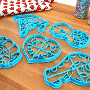 May include: A set of six blue plastic cookie cutters in various shapes, including a heart, a swirl, a donut, and a pair of glasses. The cutters are on a wooden surface.