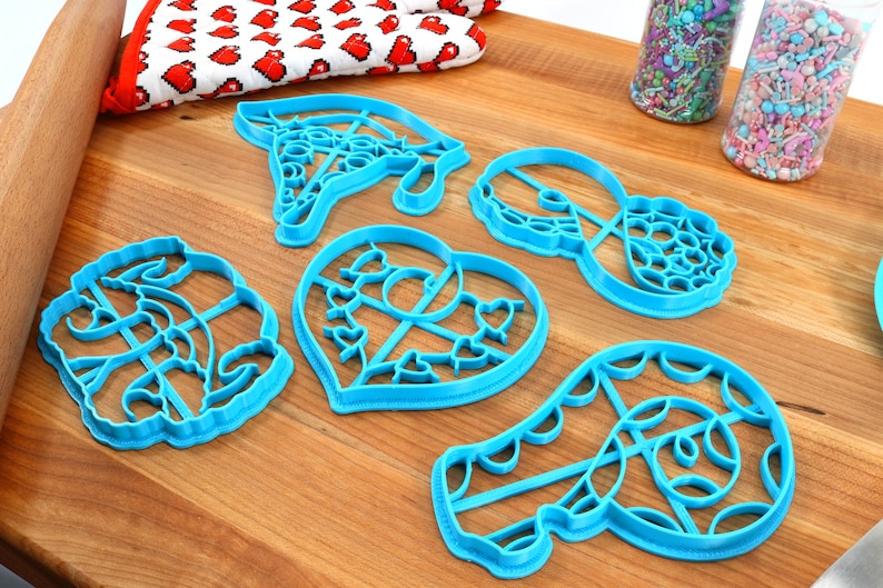 May include: A set of six blue plastic cookie cutters in various shapes, including a heart, a swirl, a donut, and a pair of glasses. The cutters are on a wooden surface.