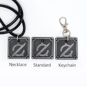 Pu&ograve; includere: Tre ciondoli color argento con un design stilizzato della lettera Z. I ciondoli sono mostrati su una collana con cordoncino nero, un ciondolo standard e un portachiavi.