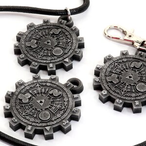 May include: Three silver-colored metal pendants with a circular design featuring a stylized Triforce symbol and other intricate details. The pendants are attached to a black cord and a silver-colored metal chain.