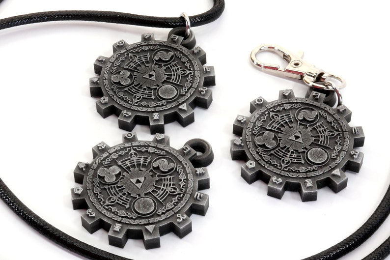 May include: Three silver-colored metal pendants with a circular design featuring a stylized Triforce symbol and other intricate details. The pendants are attached to a black cord and a silver-colored metal chain.