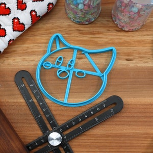 May include: A blue plastic cookie cutter shaped like a cat's face with large eyes and a wide mouth. The cutter is on a wooden surface with a ruler and a rolling pin.