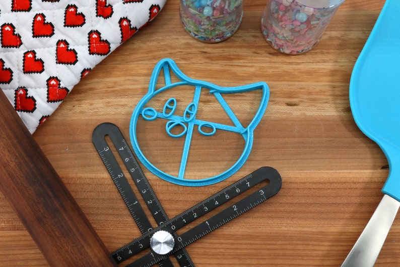 May include: A blue plastic cookie cutter shaped like a cat's face with large eyes and a wide mouth. The cutter is on a wooden surface with a ruler and a rolling pin.