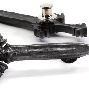 May include: Two silver-toned sword-shaped pins with intricate details. The pins have a black finish and a decorative design. The pins are attached to a white background.