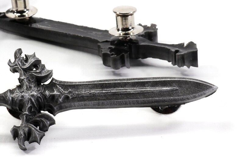 May include: Two silver-toned sword-shaped pins with intricate details. The pins have a black finish and a decorative design. The pins are attached to a white background.