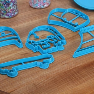 May include: A set of five blue plastic cookie cutters shaped like a skateboard, a skull, a hat, a surfboard, and a vest.