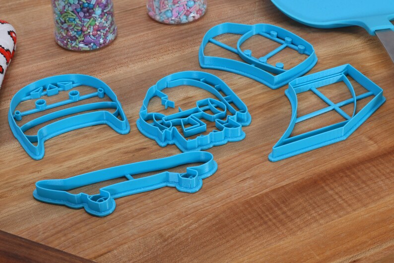 May include: A set of five blue plastic cookie cutters shaped like a skateboard, a skull, a hat, a surfboard, and a vest.
