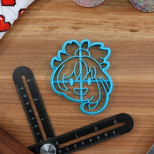May include: A blue plastic cookie cutter shaped like a cartoon character with a smiling face and long hair. The cutter is on a wooden surface.