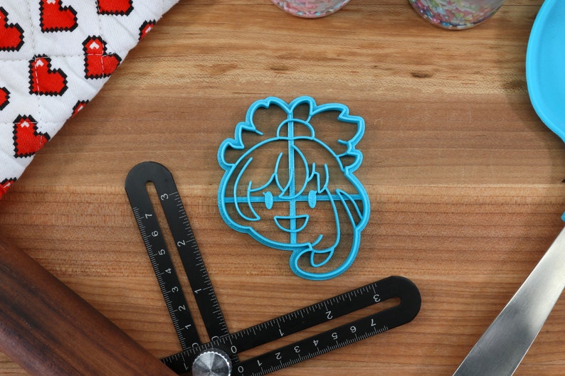 May include: A blue plastic cookie cutter shaped like a cartoon character with a smiling face and long hair. The cutter is on a wooden surface.