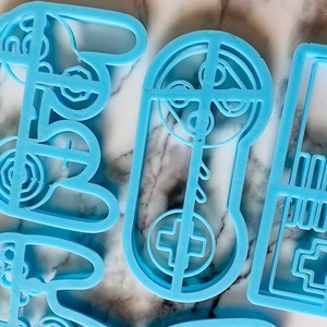 May include: Four blue plastic cookie cutters shaped like a video game controller, a joystick, and a game cartridge. The cutters are on a white marble surface.