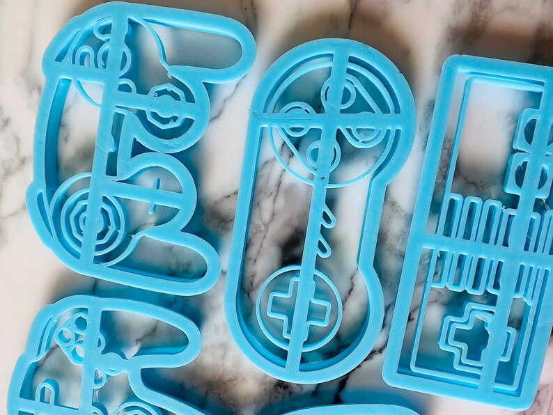 May include: Four blue plastic cookie cutters shaped like a video game controller, a joystick, and a game cartridge. The cutters are on a white marble surface.