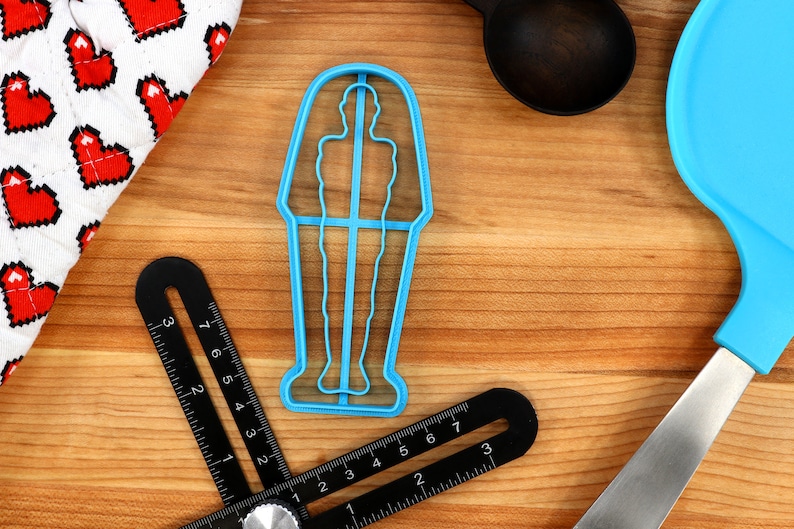 May include: A blue plastic cookie cutter shaped like a standing person in a sarcophagus. The cookie cutter is on a wooden surface.